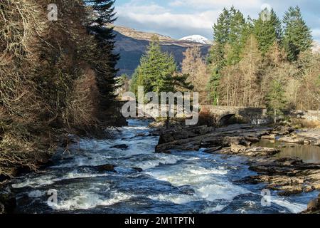 Die Wasserfälle von Dochart blicken flussabwärts in Richtung der Brücke, die weißes Wasser, Felsen, die Brücke, Kiefern und Berge im Hintergrund zeigt. Stockfoto