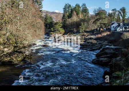 Die Wasserfälle von Dochart blicken flussabwärts in Richtung der Brücke, die weißes Wasser, Felsen, die Brücke, Kiefern, das Gasthaus und Berge im Hintergrund zeigt. Stockfoto