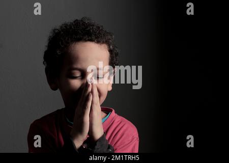 Ein kleiner Junge betet zu Gott mit Händen zusammen mit einem schwarzen Hintergrundfoto Stockfoto