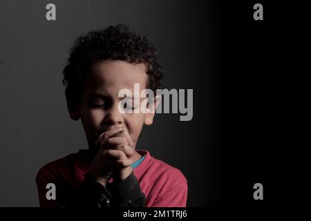 Ein kleiner Junge betet zu Gott mit Händen zusammen mit einem schwarzen Hintergrundfoto Stockfoto