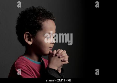 Ein kleiner Junge betet zu Gott mit Händen zusammen mit einem schwarzen Hintergrundfoto Stockfoto