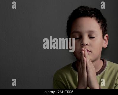 Ein kleiner Junge betet zu Gott mit Händen zusammen mit einem schwarzen Hintergrundfoto Stockfoto