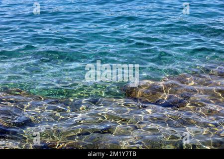 Die durchsichtige Oberfläche des Meeres mit den Steinen auf dem Grund. Felsiger Strand, türkisfarbenes Wasser als Hintergrund Stockfoto