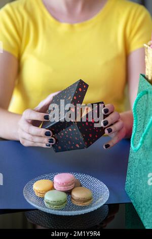 Nahaufnahme der manikürten Hände einer jungen weißen Frau mit gelbem Oberteil, während sie sich hinsetzt und eine kleine Geschenkbox öffnet, die von Geschäften umgeben ist Stockfoto