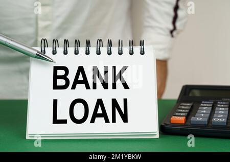 Geschäftskonzept. Auf einer grünen Oberfläche befindet sich ein Taschenrechner. Ein Mann zeigt mit einem Stift auf ein Notizbuch mit der Aufschrift „Bank Loan“ Stockfoto