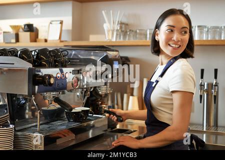 Ein lächelndes asiatisches Barista-Mädchen macht Cappuccino mit einer Kaffeemaschine, steht hinter der Theke im Café Stockfoto