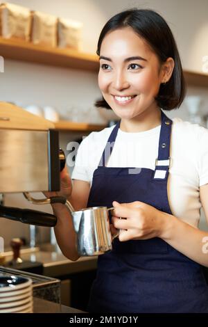 Vertikale Aufnahme eines Baristas, eines asiatischen Mädchens, das Milch für Cappuccino aufschäumt, Latte für den Gast zubereitet, eine blaue Schürze trägt, glücklich lächelt, im Café arbeitet Stockfoto