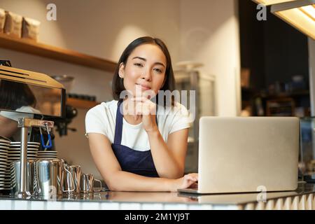 Porträt einer lächelnden koreanischen Frau, eines Baristas im Café, der mit einem Laptop an der Theke steht, lächelt und selbstbewusst aussieht, eine selbstständige Frau Stockfoto