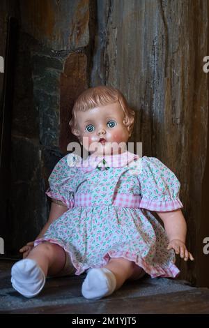 Antike Puppe in einem weißen Kleid mit pinkfarbenen Blumen und weißen Socken, die auf einem Regal aus Stein sitzt. Stockfoto