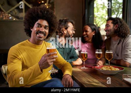 Porträt eines Mannes, der in die Kamera schaut und ein Bier hält. Freunde, die Spaß an der Bar im Hintergrund haben. Stockfoto