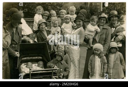 Originale Postkarte aus der Zeit Anfang der 1930er Jahre mit einer Gruppe von animierten, lebhaften Müttern im Freien an einem Frühlingstag mit vielen kleinen Kindern, Kleinkindern, Vorschuljahren, Babys im Arm, einige werden in den Armen ihrer Mütter getragen, Zwillinge im Kinderwagen, viele Charaktere. Viel Spaß haben, lächeln, vielleicht eine Gruppe von Müttern und Babys. Modische Glockenhüte für Mütter und Mädchen (modisch bis 1933) Mäntel mit großem Kragen, definierter Gürtel, Bild etwa 1930 / 1931 Großbritannien Stockfoto