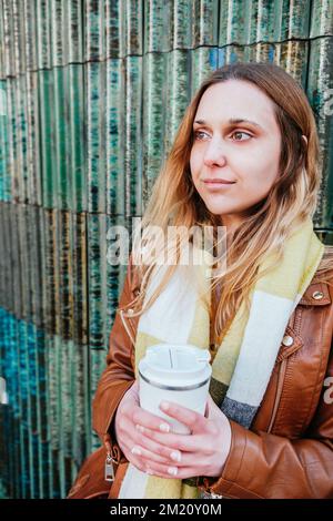 Vertikales Porträt einer attraktiven jungen Frau, die sich an die Wand lehnt. Sie hält eine Tasse Kaffee zum Mitnehmen Stockfoto