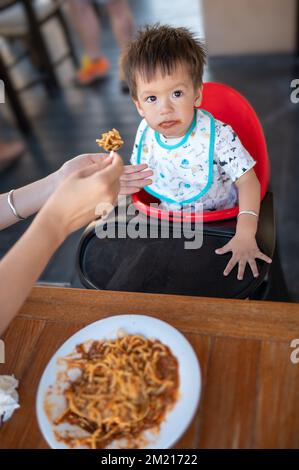 Das Kleinkind wird im Restaurant von seiner mutter gefüttert. Ein gut aussehender, eineinhalb Jahre alter, multiethnischer Junge, der mit seinem Mo im Restaurant zu Abend isst Stockfoto