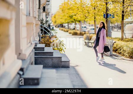 Frau Herbststadt. Eine Frau in einem rosa Kunstpelzmantel, die im Herbst an einem sonnigen Tag auf einer Straße posiert. Bäume mit gelben Blättern entlang der Straße. Stockfoto