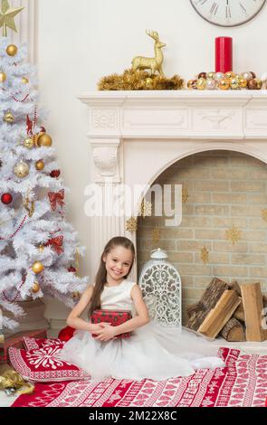 Ein kleines Mädchen in einem schönen Kleid posiert im Haus in der Nähe des Weihnachtsbaums mit einer Geschenkbox in der Hand. X-Mass und Neujahr Warten und Feiern Konzept. Stockfoto