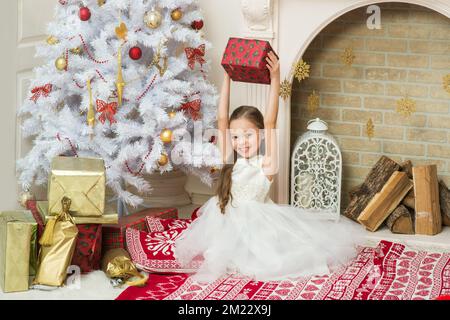 Ein kleines Mädchen in einem schönen Kleid posiert im Haus in der Nähe des Weihnachtsbaums mit einer Geschenkbox in der Hand. X-Mass und Neujahr Warten und Feiern Konzept. Stockfoto