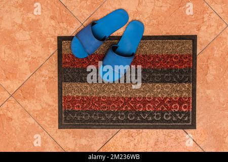 Blue homemade slippers stand on the foot carpet against the background of the floor tiles, the top view from above. Stockfoto