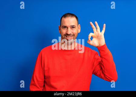 Gut aussehender, fröhlicher, bärtiger Latino-Mann in seinen 40s Jahren, der ein rotes Sweatshirt trägt und eine gute Geste isoliert vor blauem Studiohintergrund macht. Lebensstil der Menschen Stockfoto