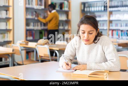 Junge Latina liest Bücher und macht Notizen in der Bibliothek Stockfoto