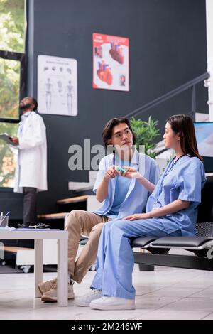 Krankenschwester, die bei asiatischen Patienten eine Diabetesrutinkontrolle mit dem Glukometer während des Termins durchführt, Assistentin bei der Blutzuckermessung am Insulintest. Junger Mann mit Kontrollbesuch in der Krankenhausrezeption Stockfoto