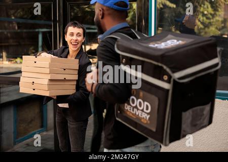 Happy customer holding pizza boxes pile, receiving order from courier outdoors, woman taking meal packages stack for colleagues. African american man delivering office lunch, food delivery service Stock Photo