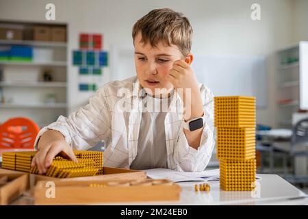 Lächelnder männlicher Junge mit Montessori-Mathematik, goldene Bank, Perlen, alternative Zählentwicklung Stockfoto