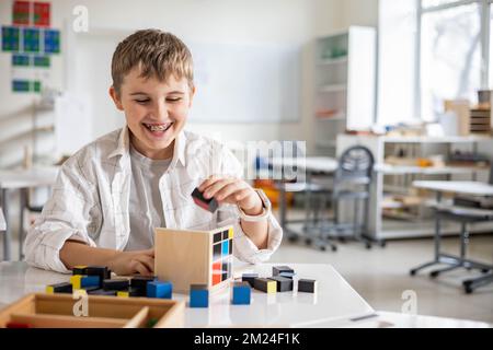 Glücklicher Junge, der Montessori-Stoff-Trinomialwürfel am Klassenschreibtisch zusammenstellt Stockfoto