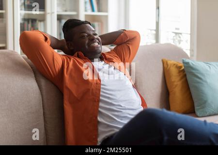 Ein glücklicher afrikanischer Millennials in lässigem Outfit, der sich auf der Couch zurücklehnt Stockfoto