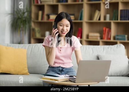 Telearbeit-Konzept. Glückliche asiatische Dame, die ihren Laptop benutzte und mit ihrem Smartphone sprach, von zu Hause aus arbeitete Stockfoto