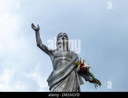 Eine alte Jesus Christus wiederauferstandene Statue mit einem mehrfarbigen Blumenstrauß in der Hand und einem blauen bewölkten Himmel im Hintergrund Stockfoto