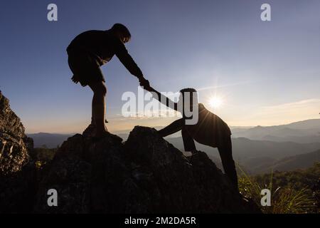 Silhouette der helfenden Hand zwischen zwei Kletterer. Paar Wandern helfen einander Silhouette in Bergen mit Sonnenlicht. Die Männer helfen, die Leute hochzuziehen f Stockfoto