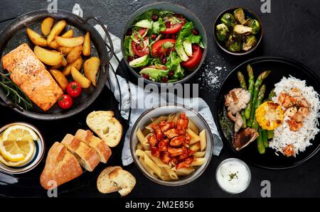 Verschiedene Hauptgerichte. Verschiedene gesunde Hauptgerichte, Fleisch- und Fischgerichte, Pasta, Salate, Saucen, Brot und Gemüse auf dunklem Hintergrund. Bis Stockfoto