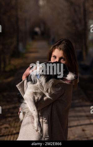 Eine weiße Frau, die einen Border Collie in den Armen hält, während sie im Herbstpark spaziert. Porträt eines Mädchens mit Hund. Stockfoto