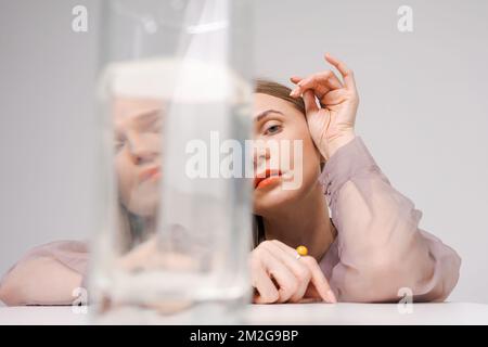 Eine schöne Frau, die durch Glaswasser schaut, blickt in die Kamera, helle orangefarbene Lippen, auf einem hellen Hintergrund im Studio. Das Konzept des emotionalen Zustands Stockfoto