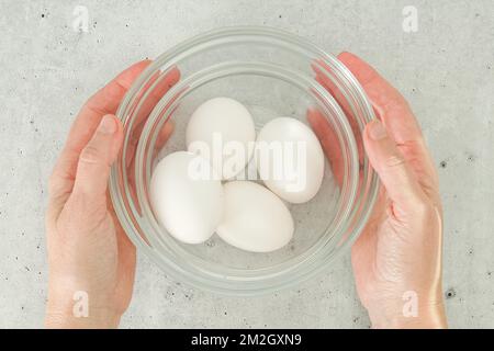 Weiße Eier in einer Glasschüssel in Frauenhänden auf hellgrauem Hintergrund direkt von oben, Kopierbereich Stockfoto
