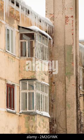 Vereiste, kleine Eiszapfen hängen im Frühling oder Winter am Dachrand. Wand eines alten Backsteinhauses mit Fenstern. Große Eiszapfen in glatten, schönen Reihen. Bewölkter Wintertag, weiches Licht. Stockfoto