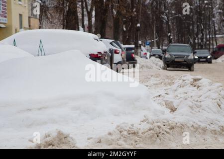 Ein großer Schneewetter vor der Kulisse einer Straße mit Autos. Auf der Straße liegt weißer Schnee in hohen Haufen. Urbane Winterlandschaft. Bewölkter Wintertag, weiches Licht. Stockfoto