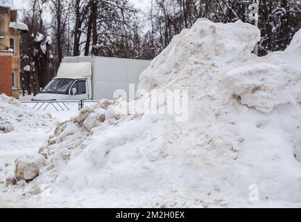 Eine riesige, schmutzige Schneeverwehung vor dem Hintergrund einer Straße mit Autos. Auf der Straße liegt weißer Schnee in hohen Haufen. Urbane Winterlandschaft. Bewölkter Wintertag, weiches Licht. Stockfoto