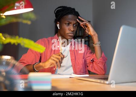 Ein junger afrikanischer Freiberufler, der sich der monotonen Aufgaben müde fühlt und mit verärgertem Gesicht auf ein Notebook schaut Stockfoto