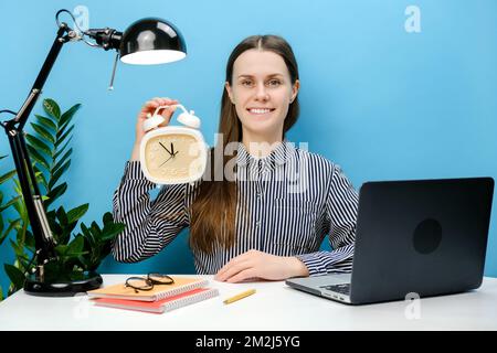 Portrait of cheerful successful employee business woman in casual shirt sit work at office desk with laptop holding in hands clock, isolated on blue s Stockfoto