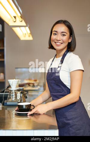 Vertikale Aufnahme einer lächelnden asiatischen Barista, trägt Uniform, macht Kaffee, steht neben der Theke mit Tasse, arbeitet im Café. Stockfoto