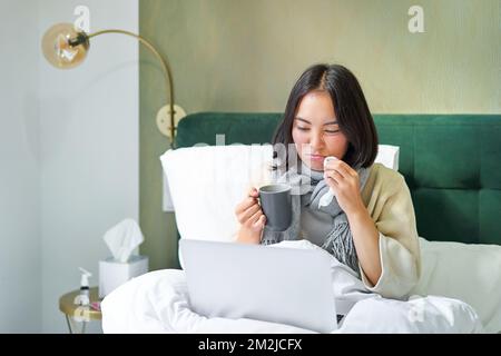 Hardworking asian girl staying in bed with cold, catching flu but working from home with laptop, studying while feeling sick Stock Photo