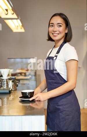 Vertical shot of smiling asian girl barista, wearing uniform, making coffee, standing near counter with cup, working in cafe Stock Photo