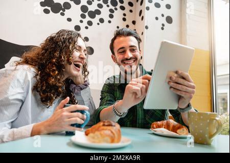 Ein junges Paar, das lächelt und auf ein Tablet schaut. Leute sitzen am Tisch in einem Café und frühstücken. Stockfoto