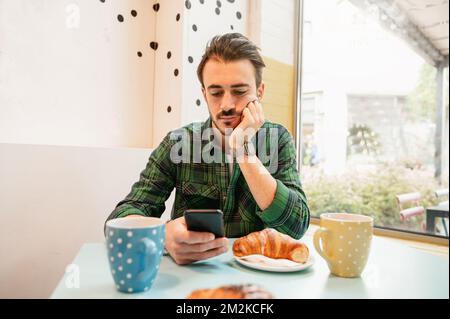 Ein bärtiger Hipstermann, der in einem Café am Tisch sitzt und frühstückt. Ein Mann, der ein Smartphone benutzt. Stockfoto