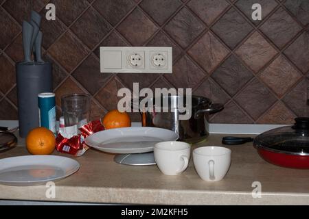 Foto vom Chaos in der Küche. Auf dem Tisch stehen Teller, Tassen, eine Orange, ein Topf, eine Pfanne Stockfoto