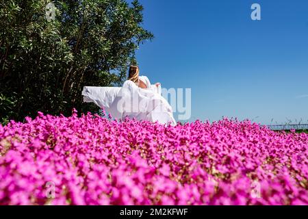 Eine schöne Frau in einem weiß fließenden langen Kleid bleiben in der Nähe eines schönen Feldes mit rosa Blumen Rückansicht Stockfoto