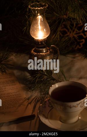 Fotografieren Sie eine brennende Kerosinlampe und einen leeren Retro-Becher und Untertasse neben einem alten Buch und einem Fichtenzweig Stockfoto