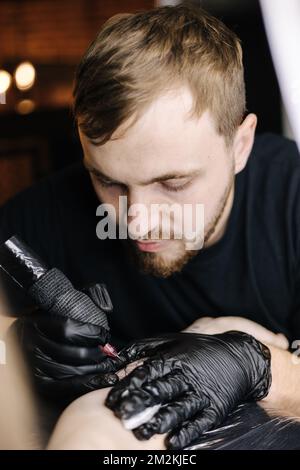 Bei der Nahaufnahme eines jungen professionellen Tätowierers wird schwarze Tinte mit einer Nadel aus einem kabellosen Tätowiergerät in die Haut eingeführt. Tattoo auf der Hand. Stockfoto