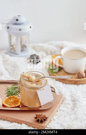 Ästhetisches Weihnachtsfrühstück. Hausgemachtes, mit Haselnuss gesalzenes Karamell in Glasgefäß mit Nachahmungsetikett zu Hause mit einer Tasse Kaffee. Stockfoto